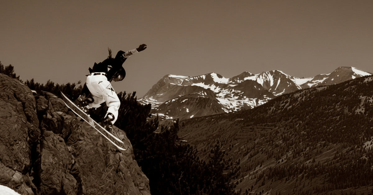 A snowboarder in mid-air performs a jump over a rocky mountain slope, wearing MHD helmet and goggles