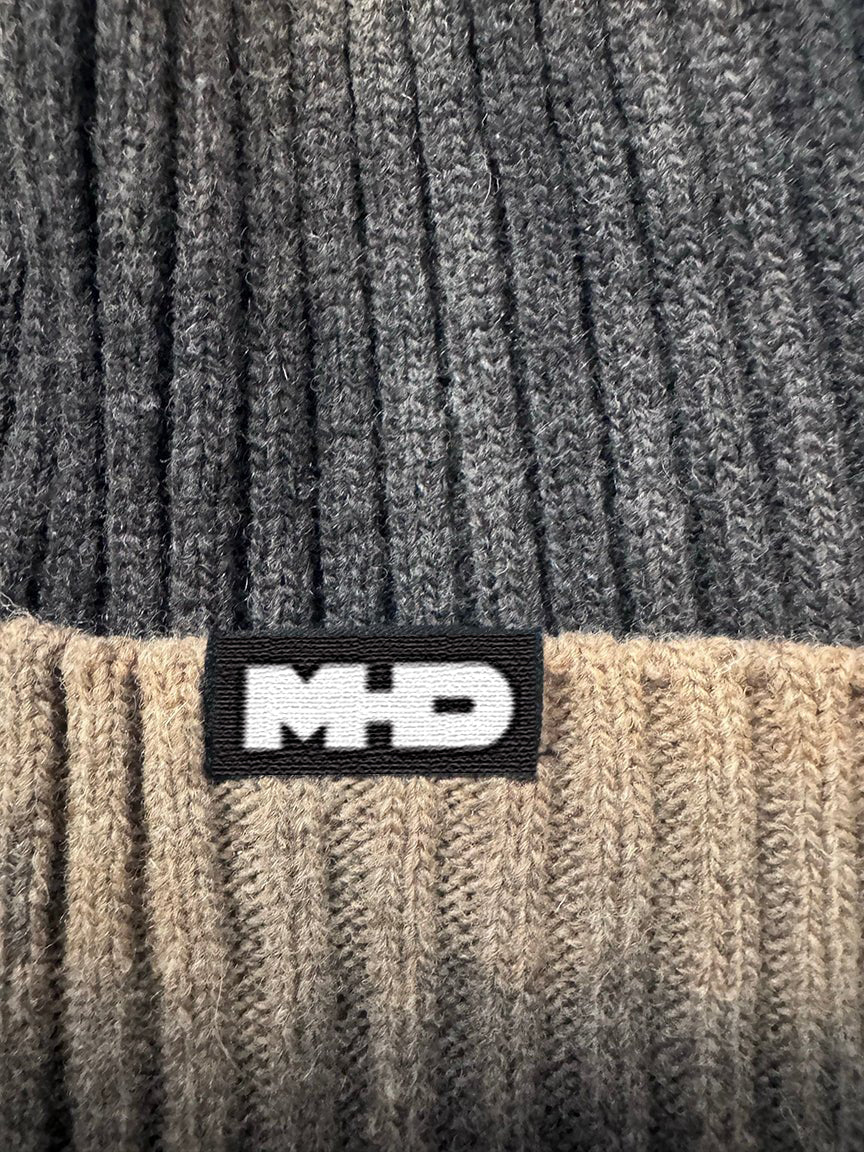 Close-up of a ribbed knit beanie in dark charcoal gray and beige, featuring a black MHD patch